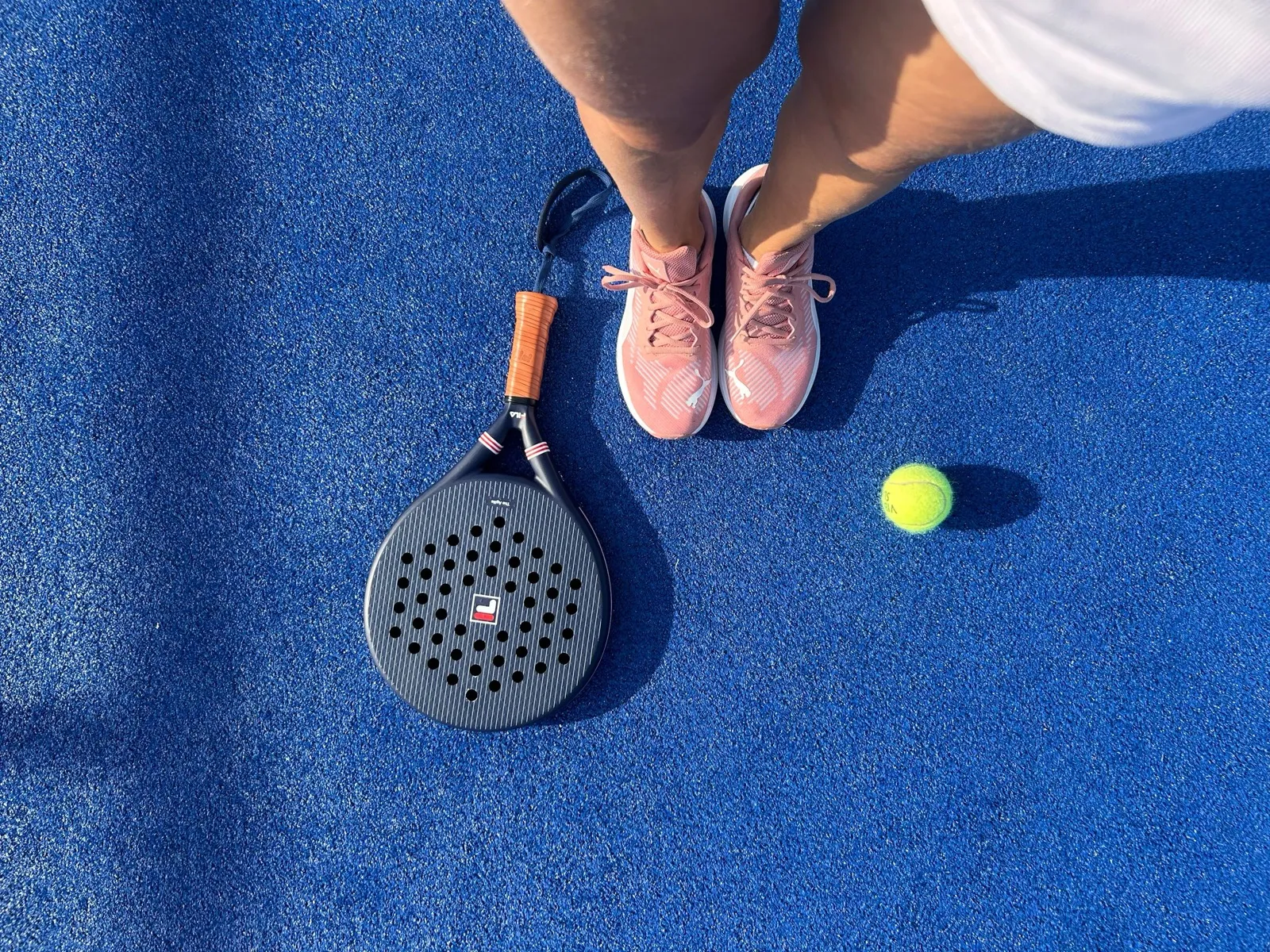 Pieds en baskets roses, raquette de padel et balle vus du dessus sur un court bleu